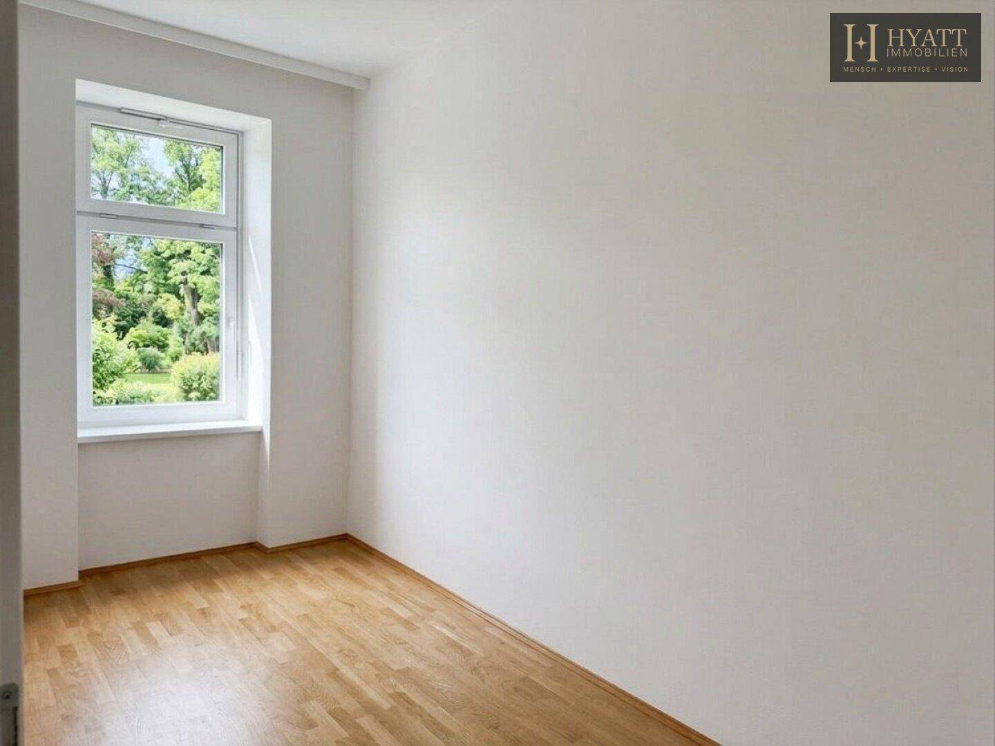 Helles, leeres Schlafzimmer mit Parkettboden und Fenster mit Blick auf grüne Bäume.