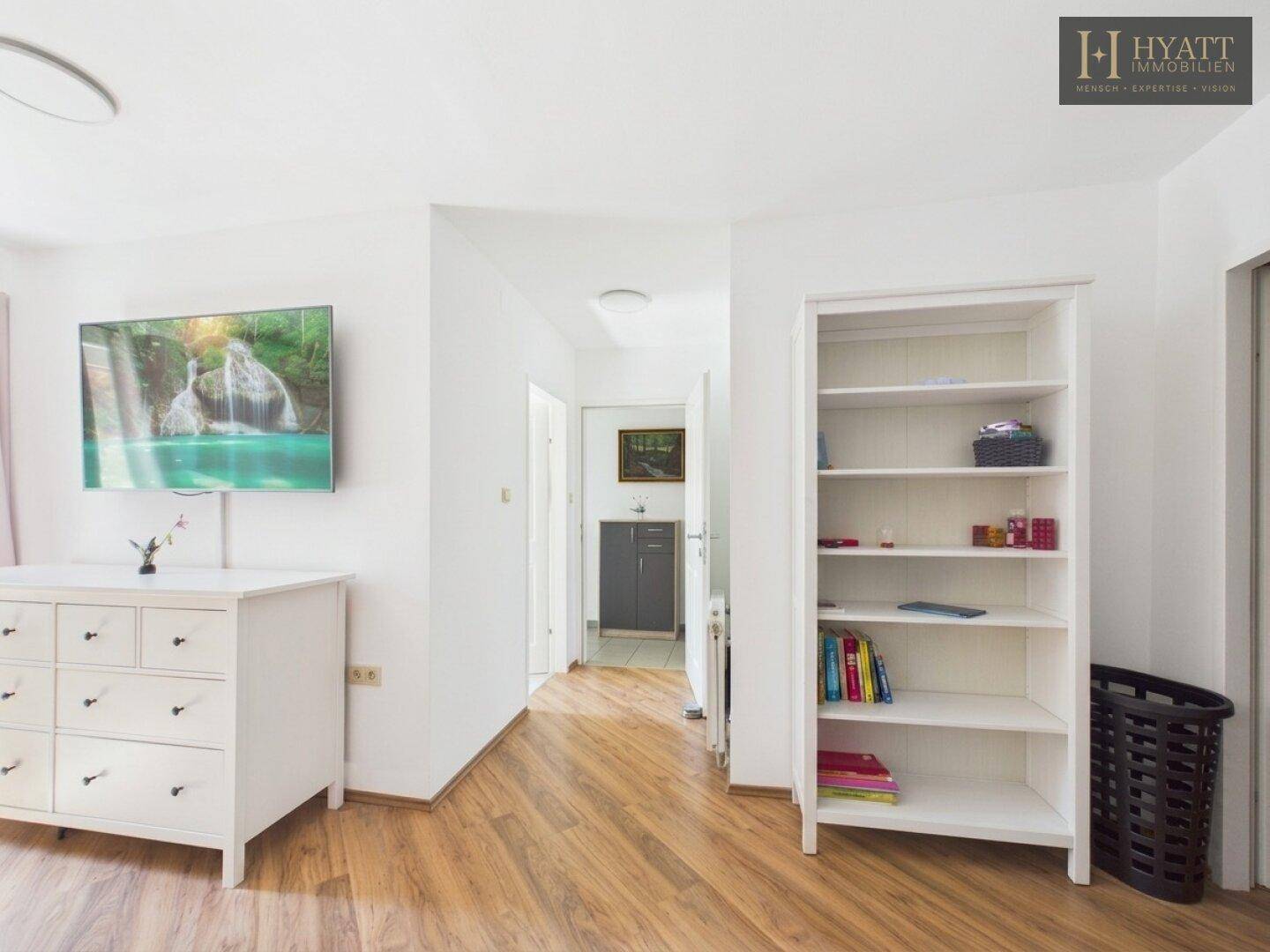 Heller Raum mit Holzboden, Kommode, Bücherregal und Fernseher.