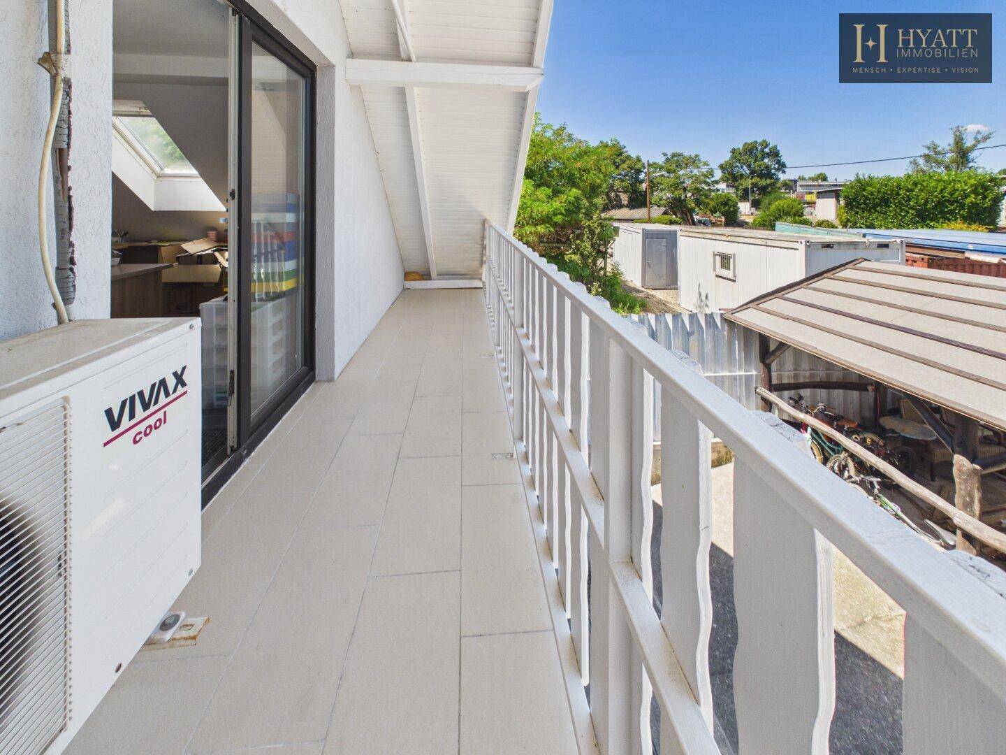 Langer Balkon mit Fliesenboden, weißem Geländer und Klimaanlage, mit Blick auf die Umgebung.