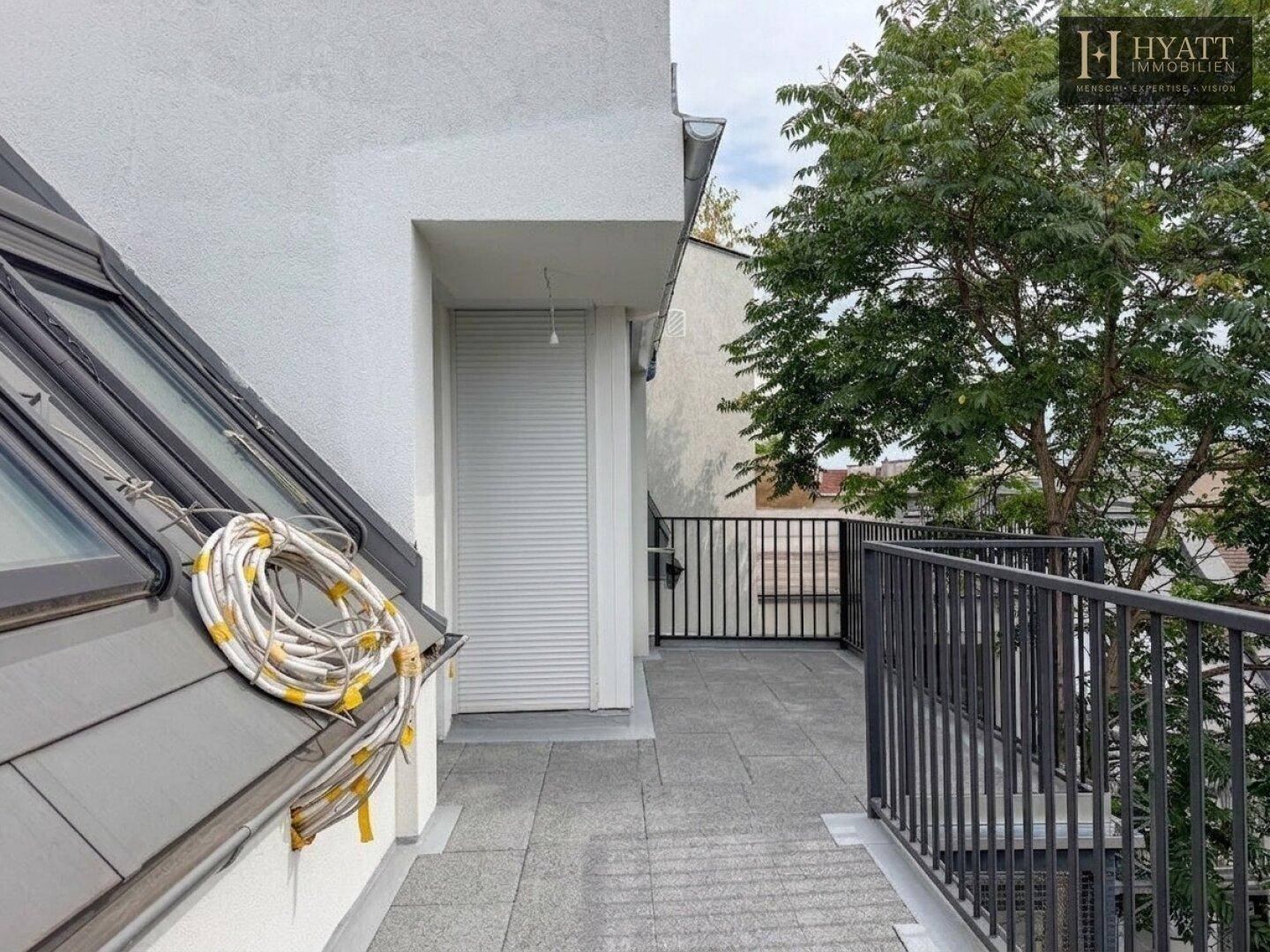 Unmöblierte Terrasse mit grauen Fliesen, schwarzem Geländer und einem Baum.