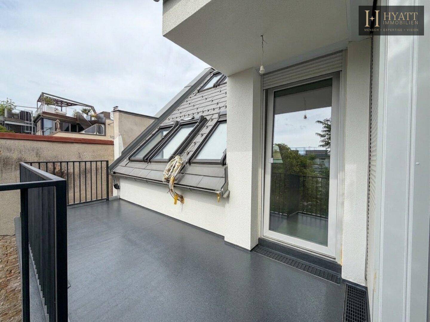 Unmöblierte Dachterrasse mit schwarzem Geländer, Dachfenstern und Blick auf die Umgebung.