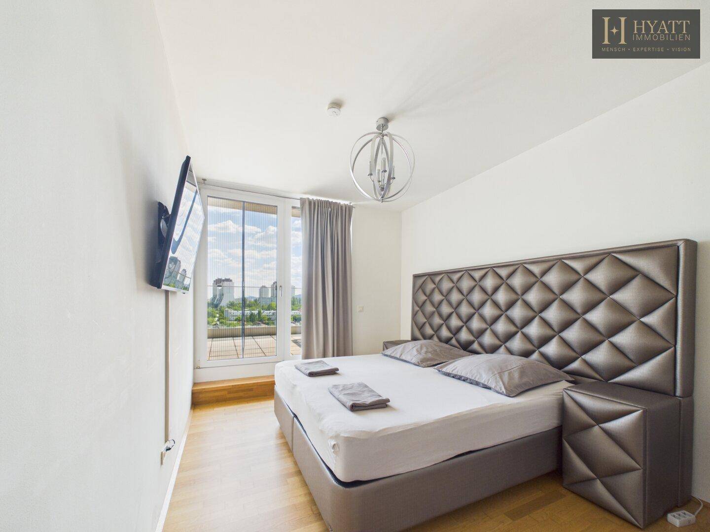 Modernes Schlafzimmer mit gepolstertem Bett, Holzboden und großen Fenstern mit Stadtblick.
