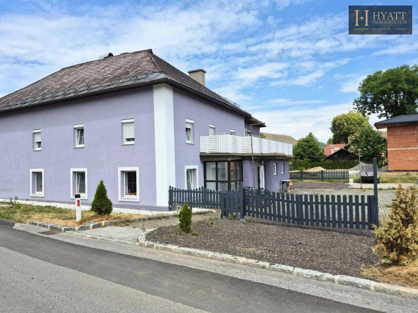 Ansicht des lila Gebäudes mit weißem Balkon und angrenzendem unbebautem Grundstück unter klarem Himmel.