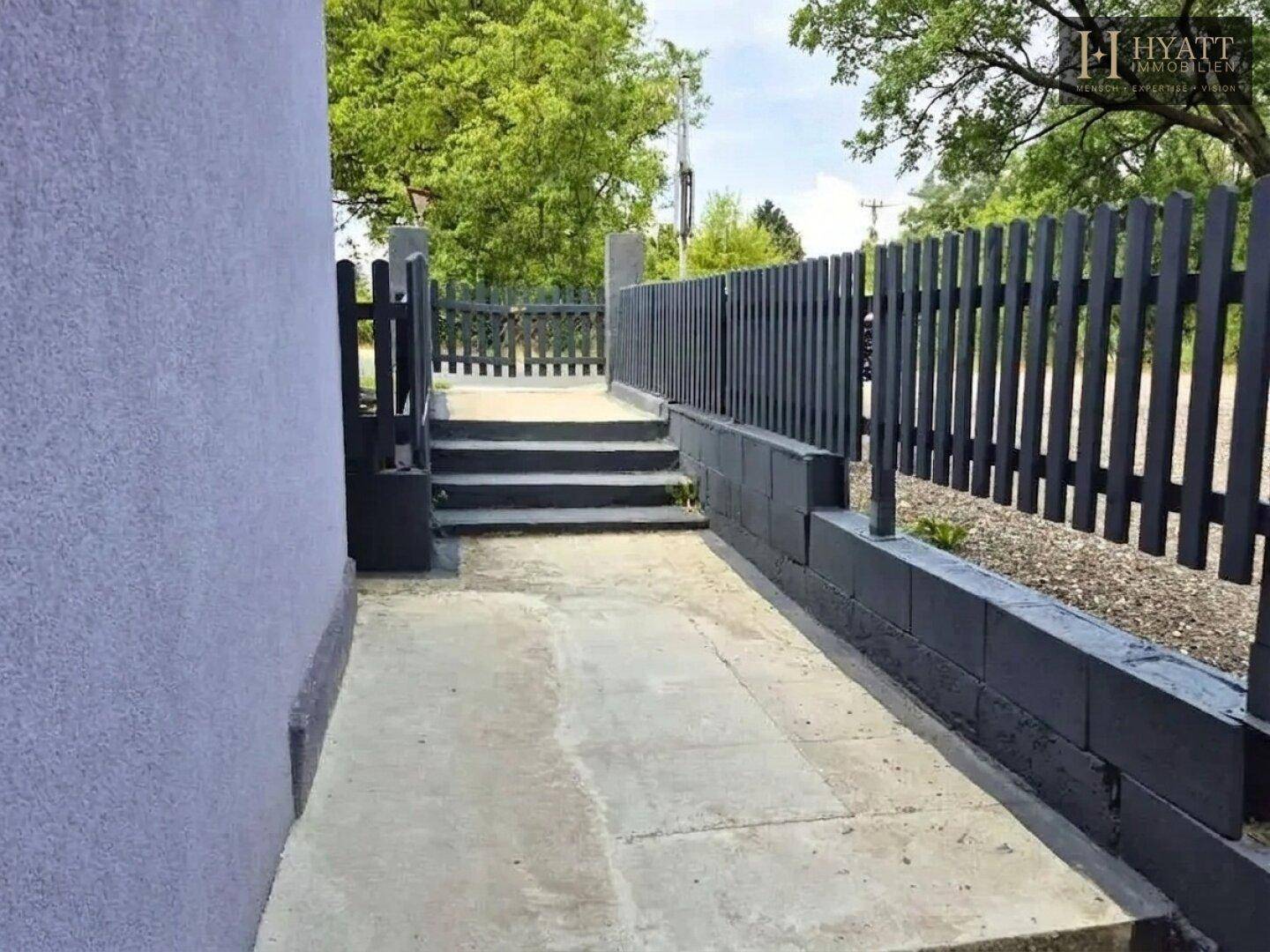 Außenweg entlang des Gebäudes mit Treppen und einem dunklen Zaun, umgeben von Grün.