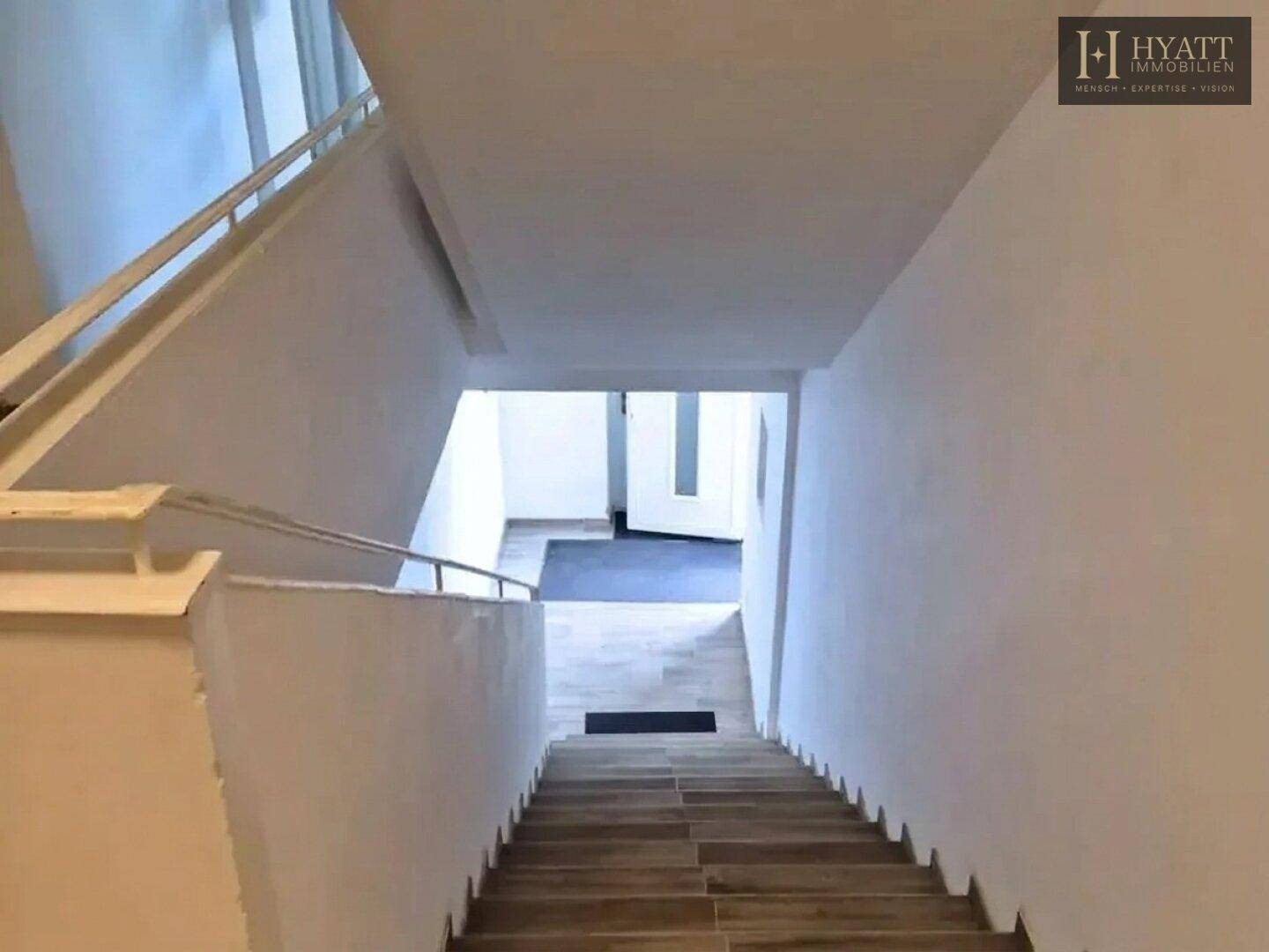 Heller Treppenbereich mit Holzstufen und weißen Wänden, der zu einem unteren Stockwerk führt.