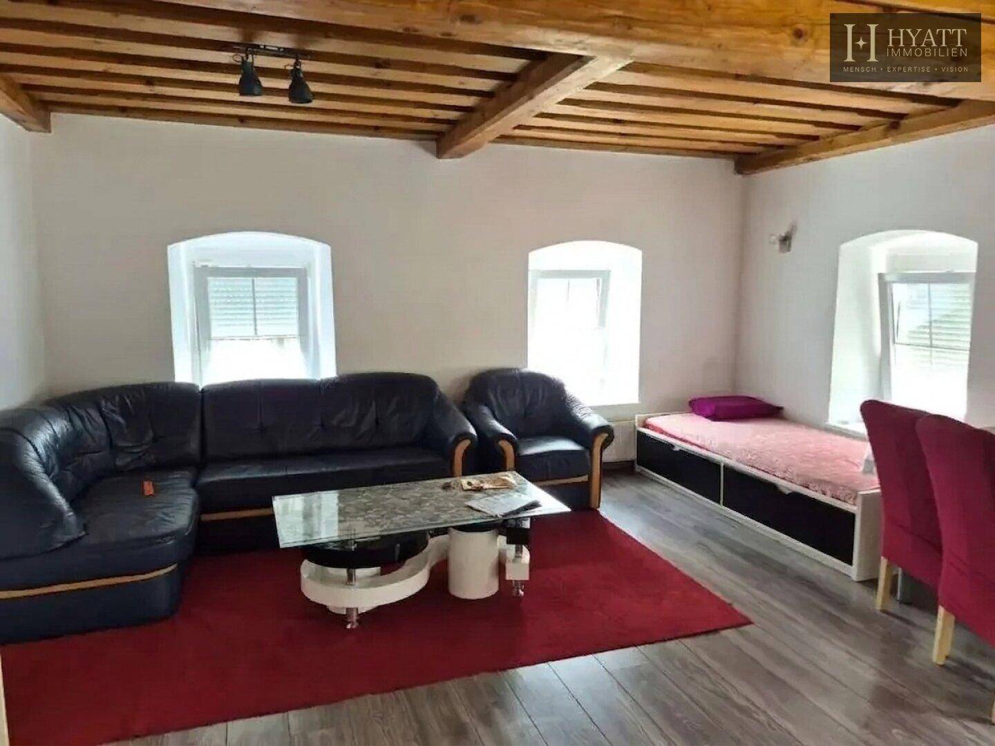 Rustikales Wohnzimmer mit schwarzem Ledersofa, Holzbalkendecke und rotem Teppich.