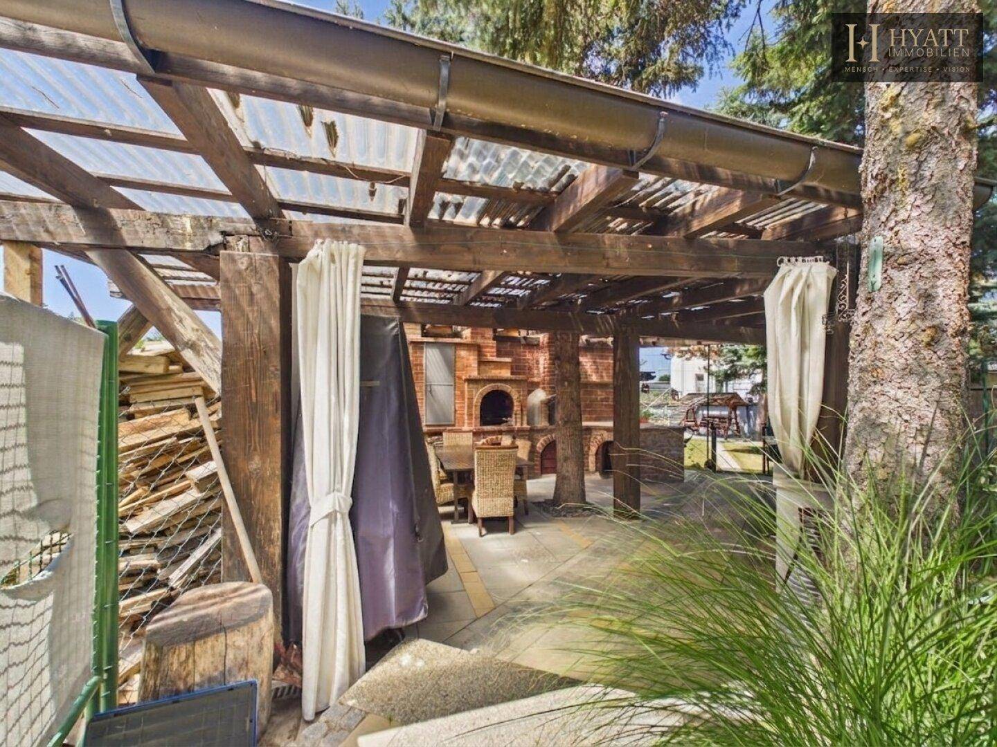 Überdachter Essbereich im Freien mit Holzpergola, gemauertem Ofen und Korbmöbeln.