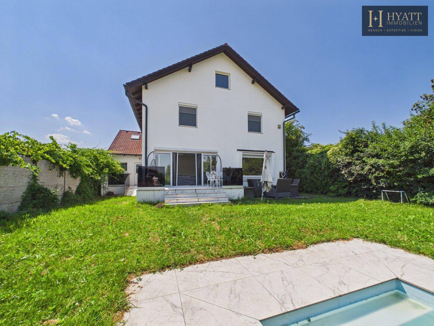 Rückansicht eines weißen Einfamilienhauses mit Garten, Rasenfläche und kleinem Pool.