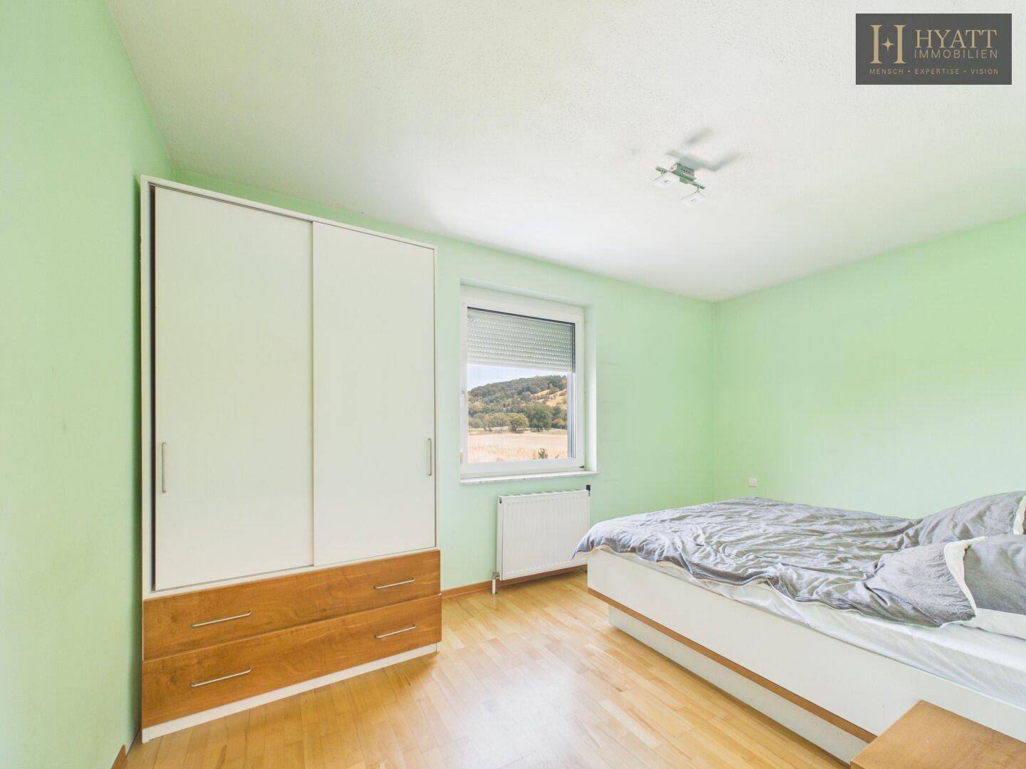 Schlafzimmer mit hellgrünen Wänden, Holzboden und einem Fenster mit Blick auf die Landschaft.
