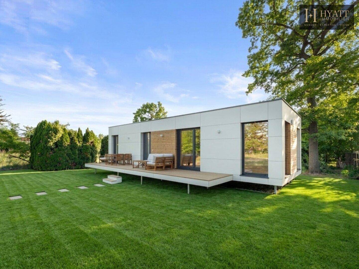 Modernes Einfamilienhaus mit weitläufigem Garten und großer Holzterrasse unter blauem Himmel.