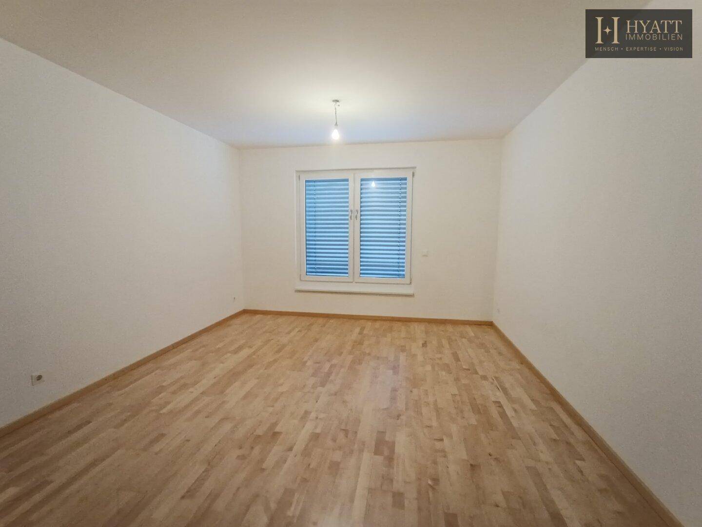 Schlichtes Zimmer mit hellem Parkettboden und einem großen Fenster, das viel Tageslicht hereinlässt.