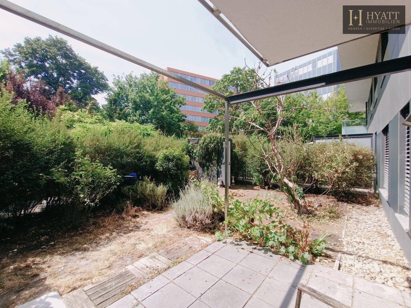 Gepflasterte Terrasse umgeben von üppigem Grün und Bäumen, mit Blick auf ein Gebäude.