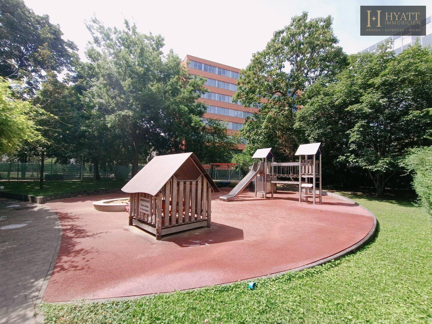 Ein großer Spielplatz mit Rutschen, Sandkasten und Spielhaus, umgeben von Bäumen und Gebäuden.