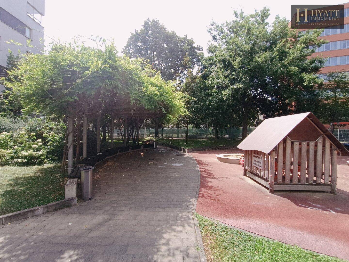 Ein schattiger Außenbereich mit überdachter Sitzgelegenheit, Sandkasten und Spielhaus, umgeben von viel Grün.