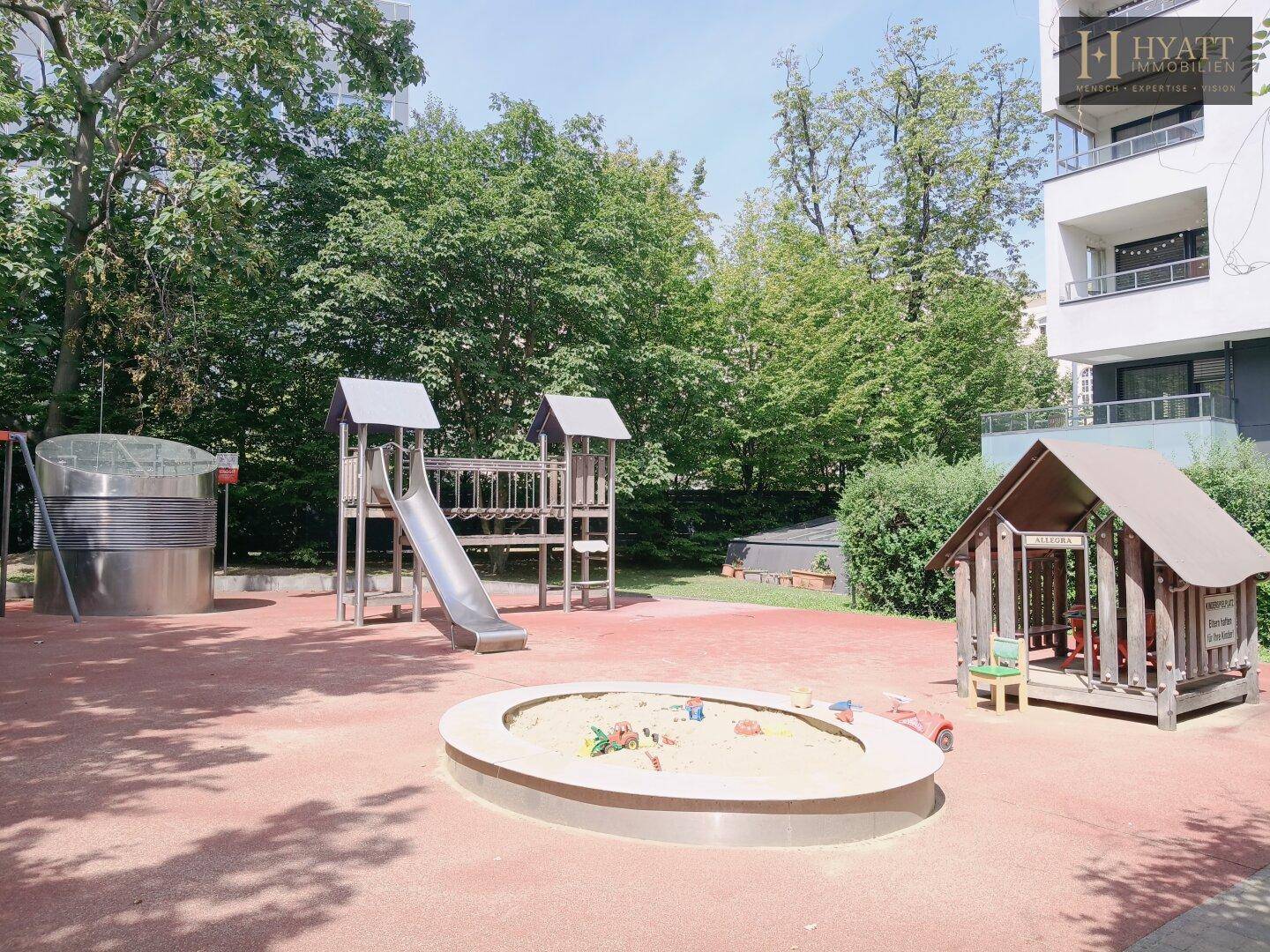 Ein gepflegter Spielplatz mit Sandkasten, Rutsche und Spielhaus vor einem modernen Wohngebäude.