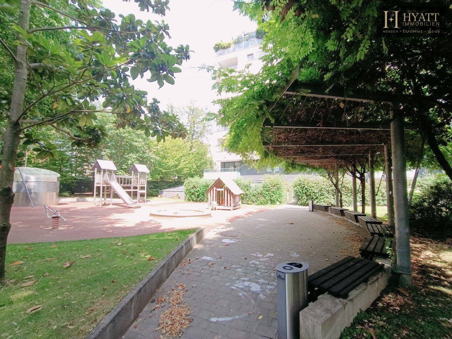 Ein idyllischer Gartenbereich mit Sitzbänken unter einer Pergola und einem Spielplatz im Hintergrund.