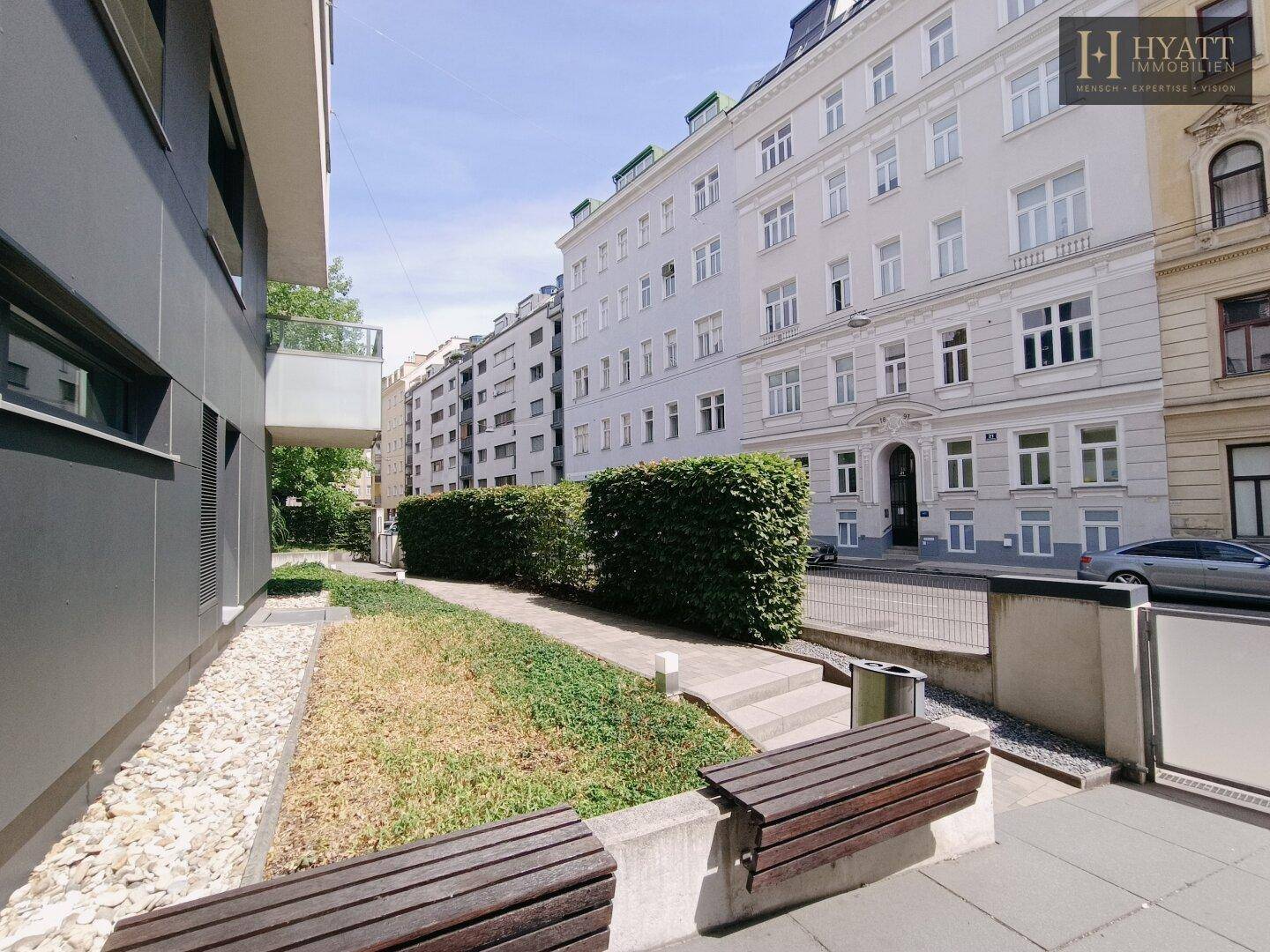 Außenansicht einer Straße mit modernen und älteren Gebäuden, gesäumt von Hecken und Sitzbänken.