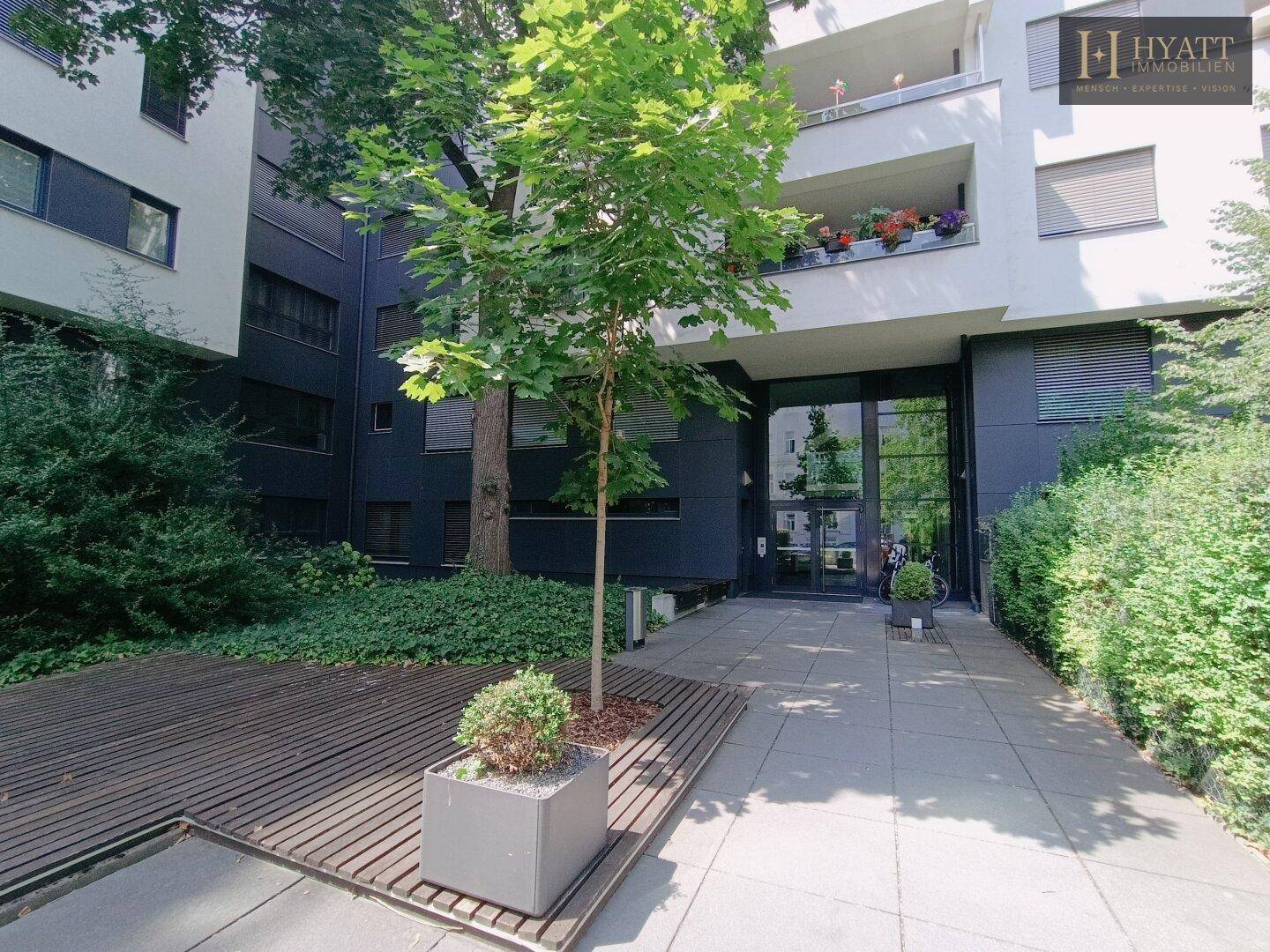 Moderner Eingangsbereich eines Gebäudes mit Glasfassade, Holzdeck und einem Baum in einem Pflanzgefäß.
