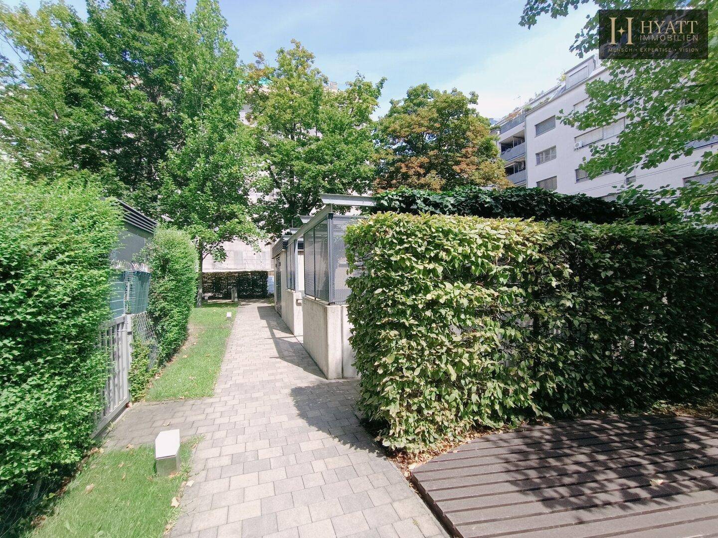 Ein gepflasterter Weg führt zwischen hohen Hecken und einem Gebäude entlang zu einer Holzterrasse.