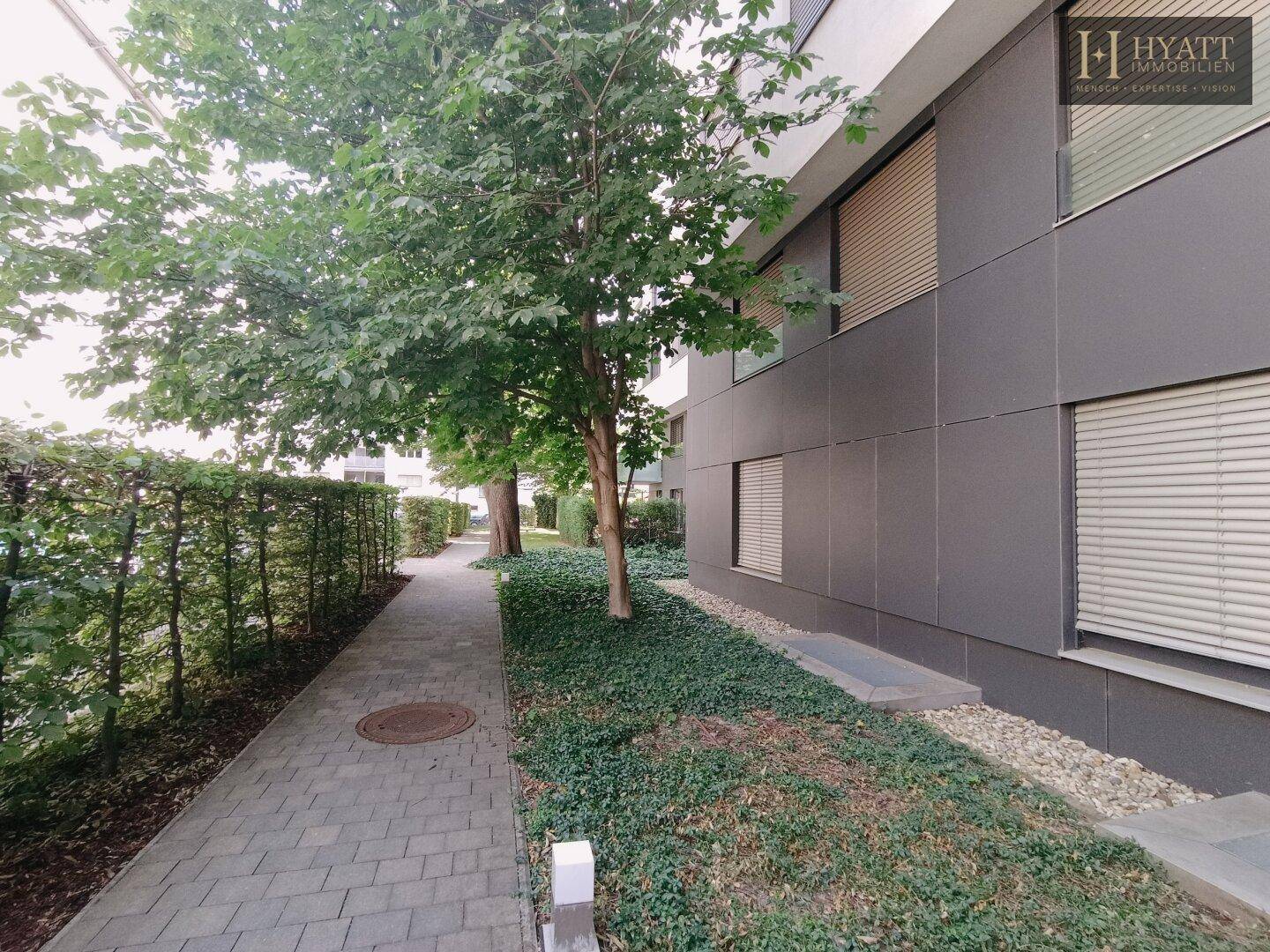 Ein gepflasterter Weg führt entlang einer modernen Gebäudefassade mit dunklen Paneelen und Hecken.