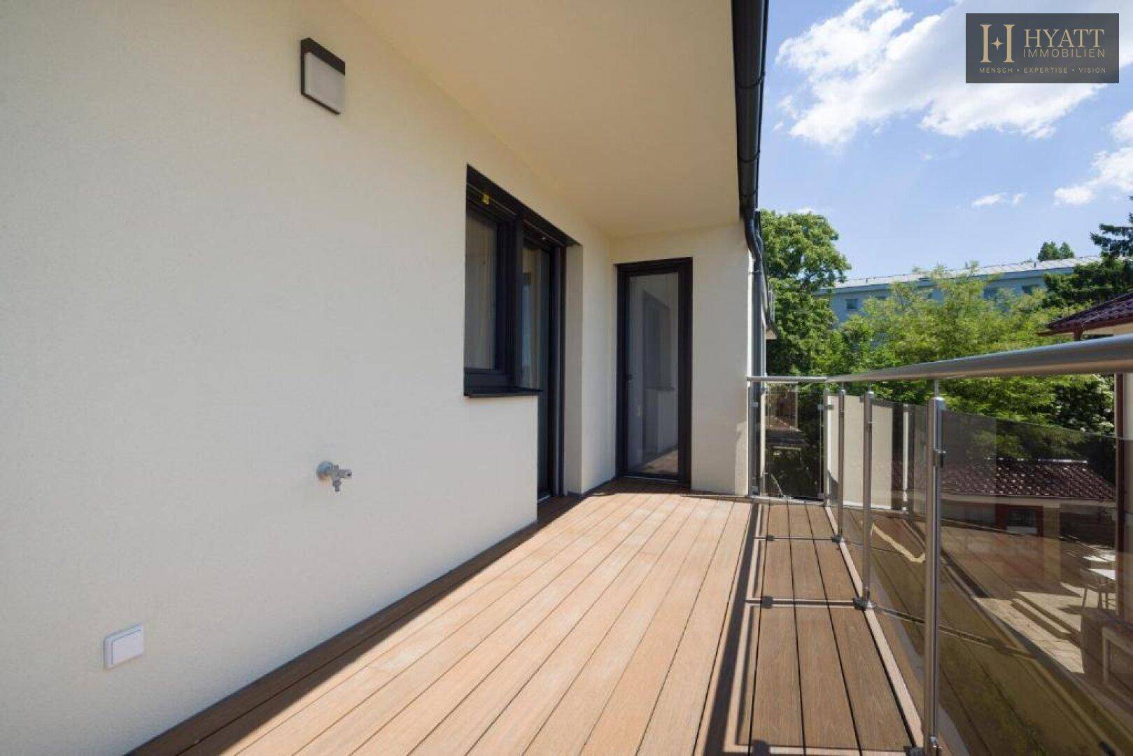 Geräumiger Balkon mit Holzbelag und Glasgeländer, umgeben von grünen Bäumen.