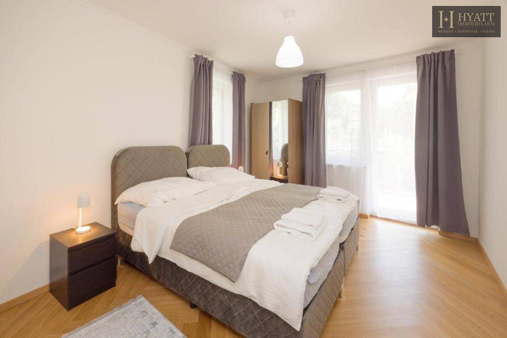Helles Schlafzimmer mit Doppelbett, Nachttisch und großen Fenstern mit Vorhängen.