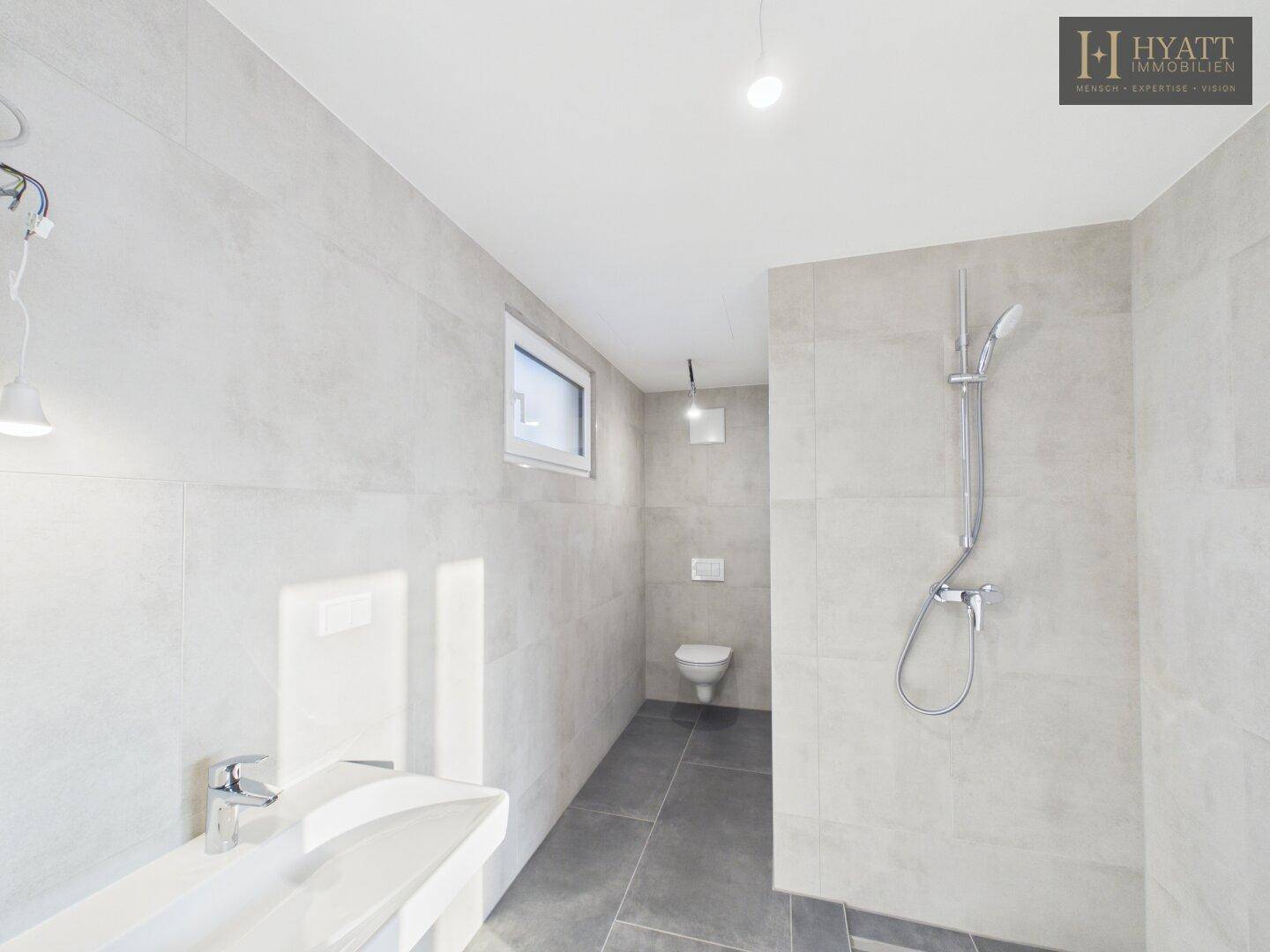 Modernes Badezimmer mit Dusche, WC und Waschbecken in hellgrauen Fliesen.