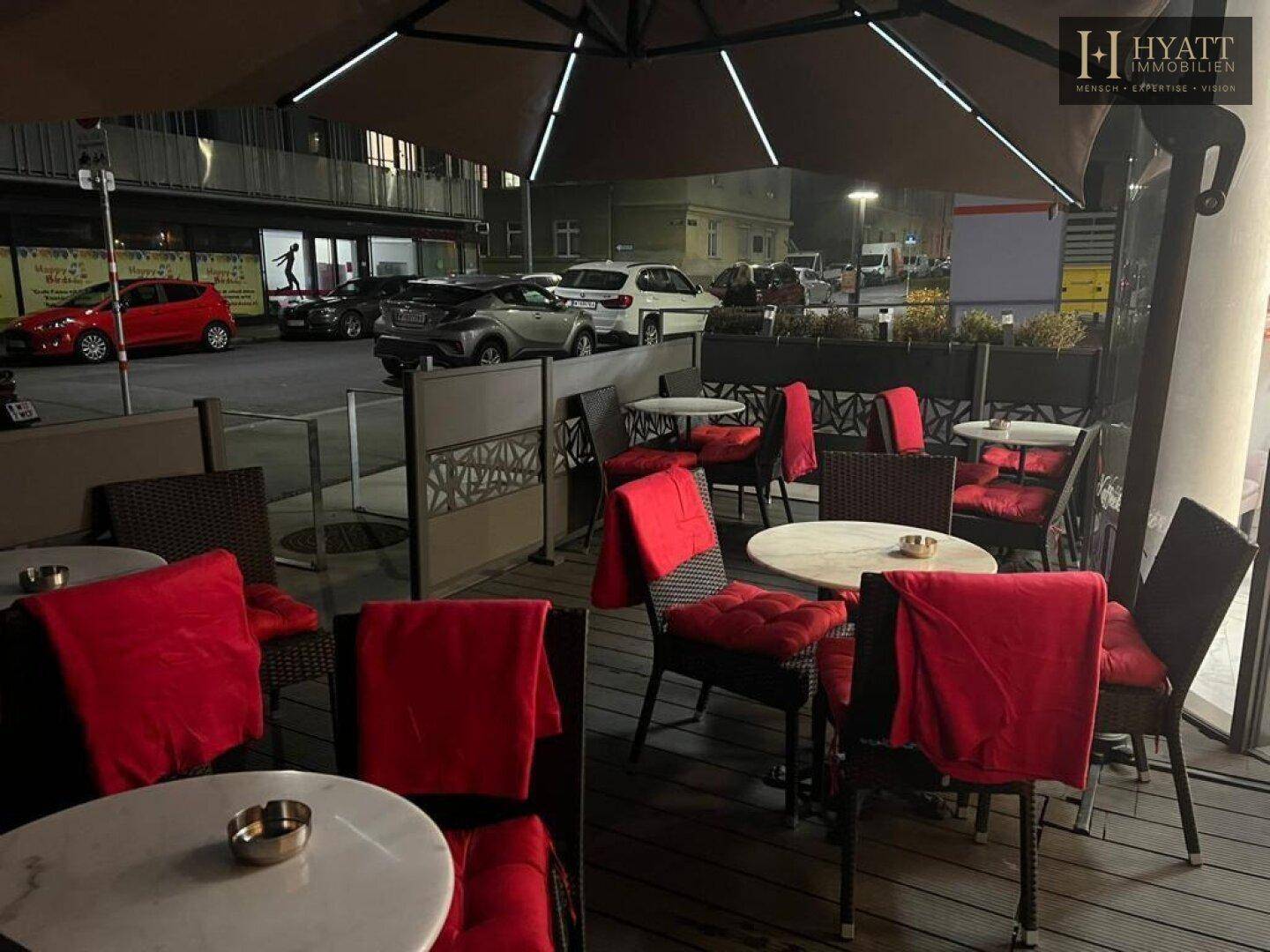 Gemütlicher Außenbereich eines Cafés bei Nacht mit roten Sitzkissen und Tischen unter einem Sonnenschirm.