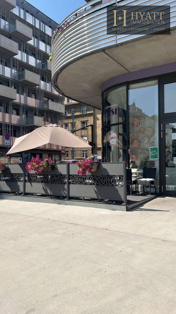 Außenbereich eines Cafés mit Sonnenschirm, Blumenkästen und Blick auf moderne Stadtgebäude.