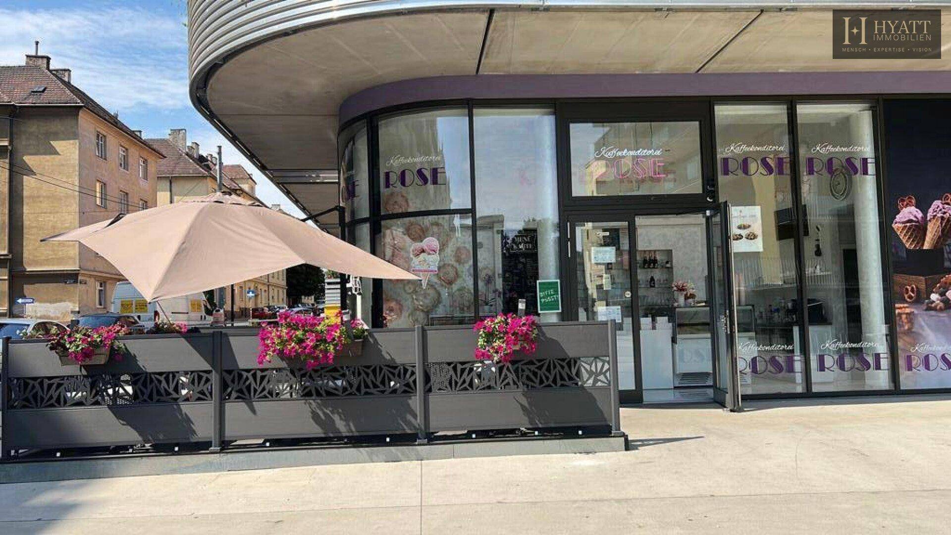 Straßenseite eines Cafés mit Außenterrasse, Sonnenschirm und Blick auf die umliegenden Gebäude.