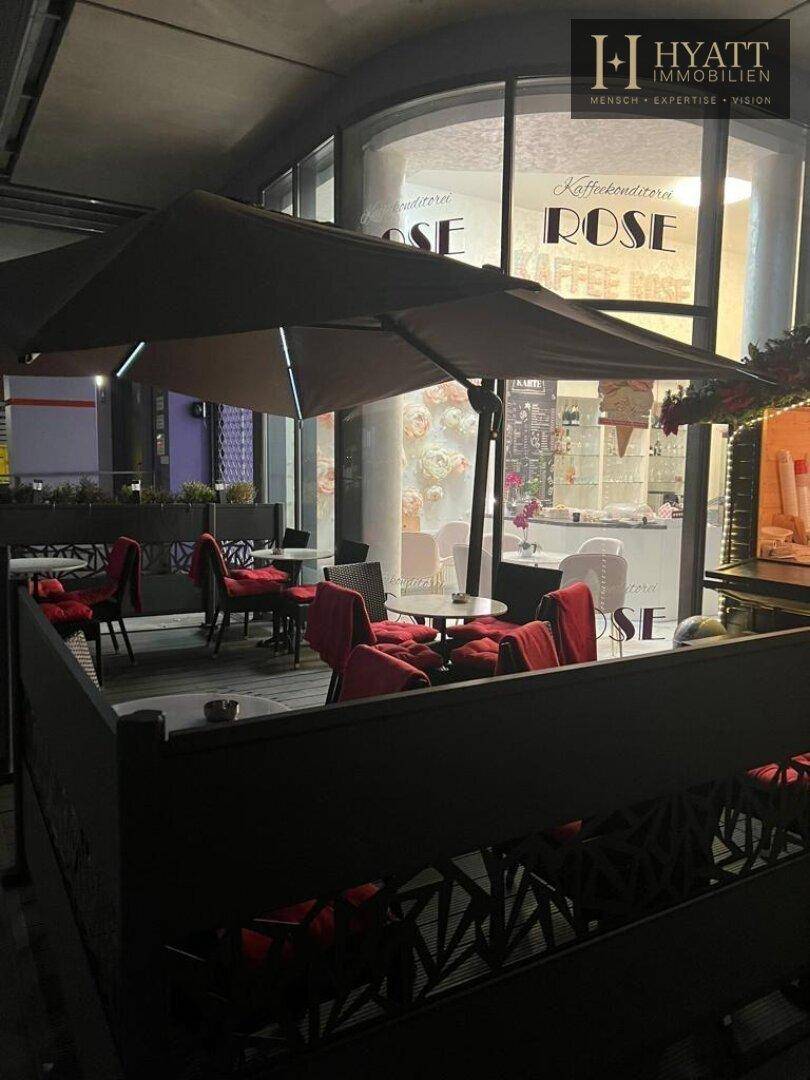Außenbereich eines Cafés bei Nacht mit Tischen, roten Stühlen und Blick auf den beleuchteten Innenraum.