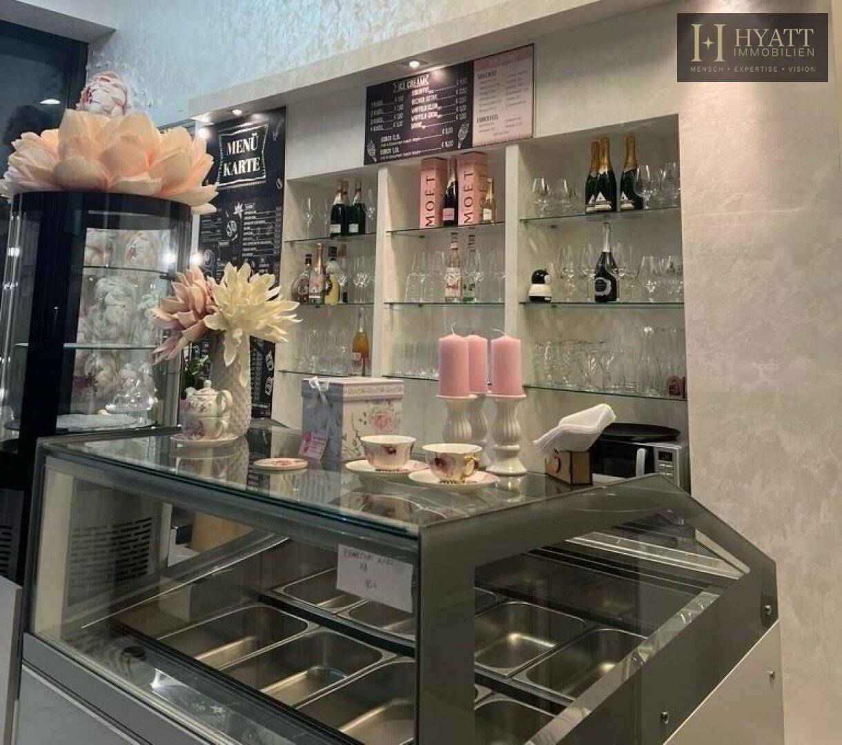 Thekenbereich eines Cafés mit Vitrine, Regalen voller Gläser und Flaschen sowie einer Menütafel.