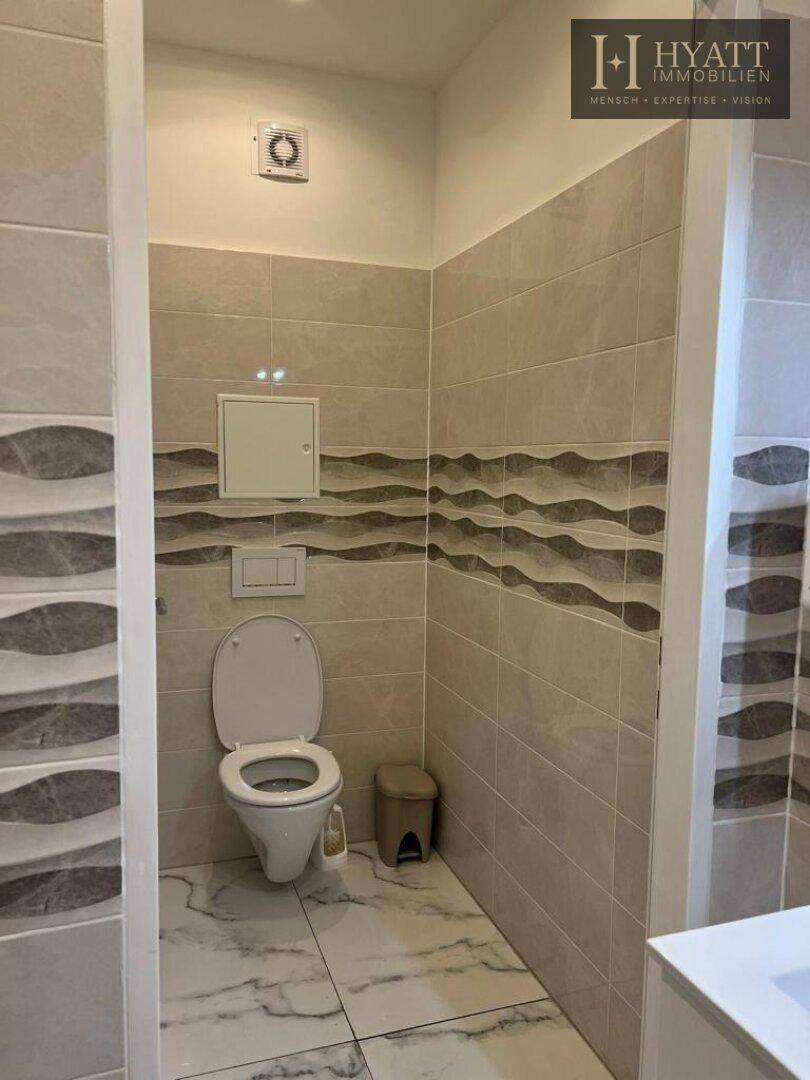 Separate Toilette mit Wand-WC, Spülkasten und Fliesen in Beige- und Grautönen.