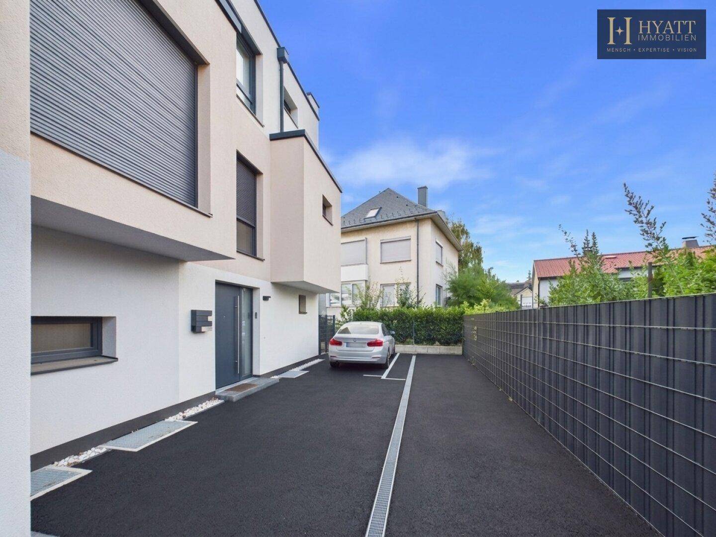 Moderne Hausfassade mit Zufahrt und Parkplatz, umgeben von einer gepflegten Hecke.