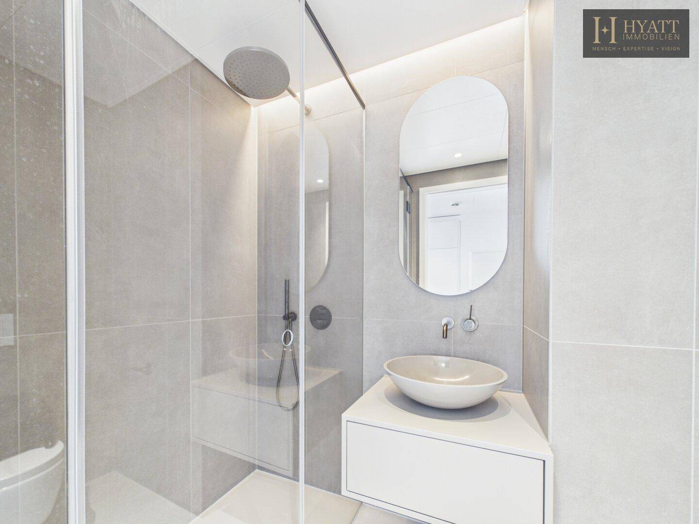 Modernes Badezimmer mit begehbarer Dusche, eleganten grauen Fliesen und ovalem Spiegel.
