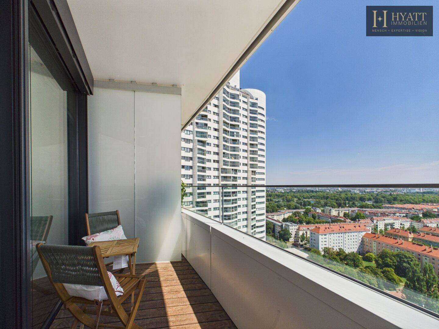 Geräumiger Balkon mit Holzboden, Sitzgelegenheiten und weitem Blick über die Stadt und Grünflächen.
