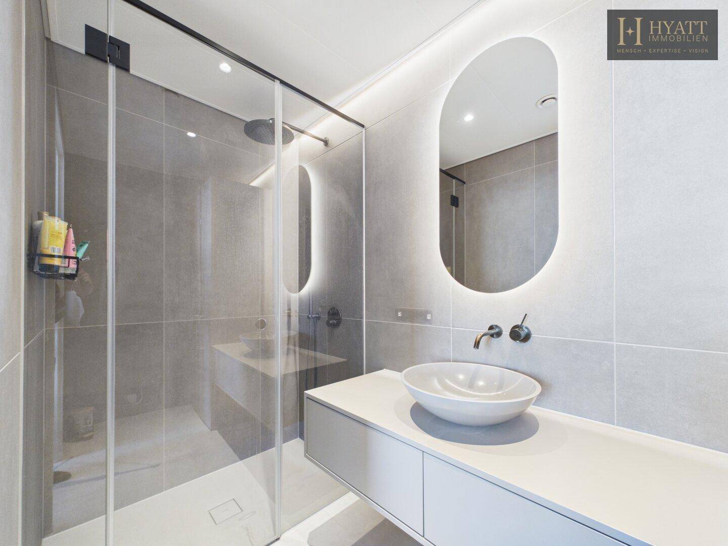 Modernes Badezimmer mit begehbarer Dusche, ovalem Spiegel und hellgrauen Fliesen.