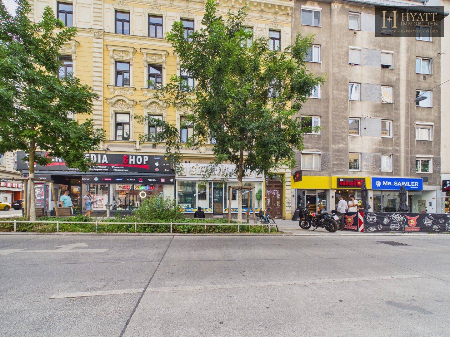 Straßenansicht mit mehreren Geschäftsfassaden, darunter "SHAM Sweets", und Bäumen am Gehweg.