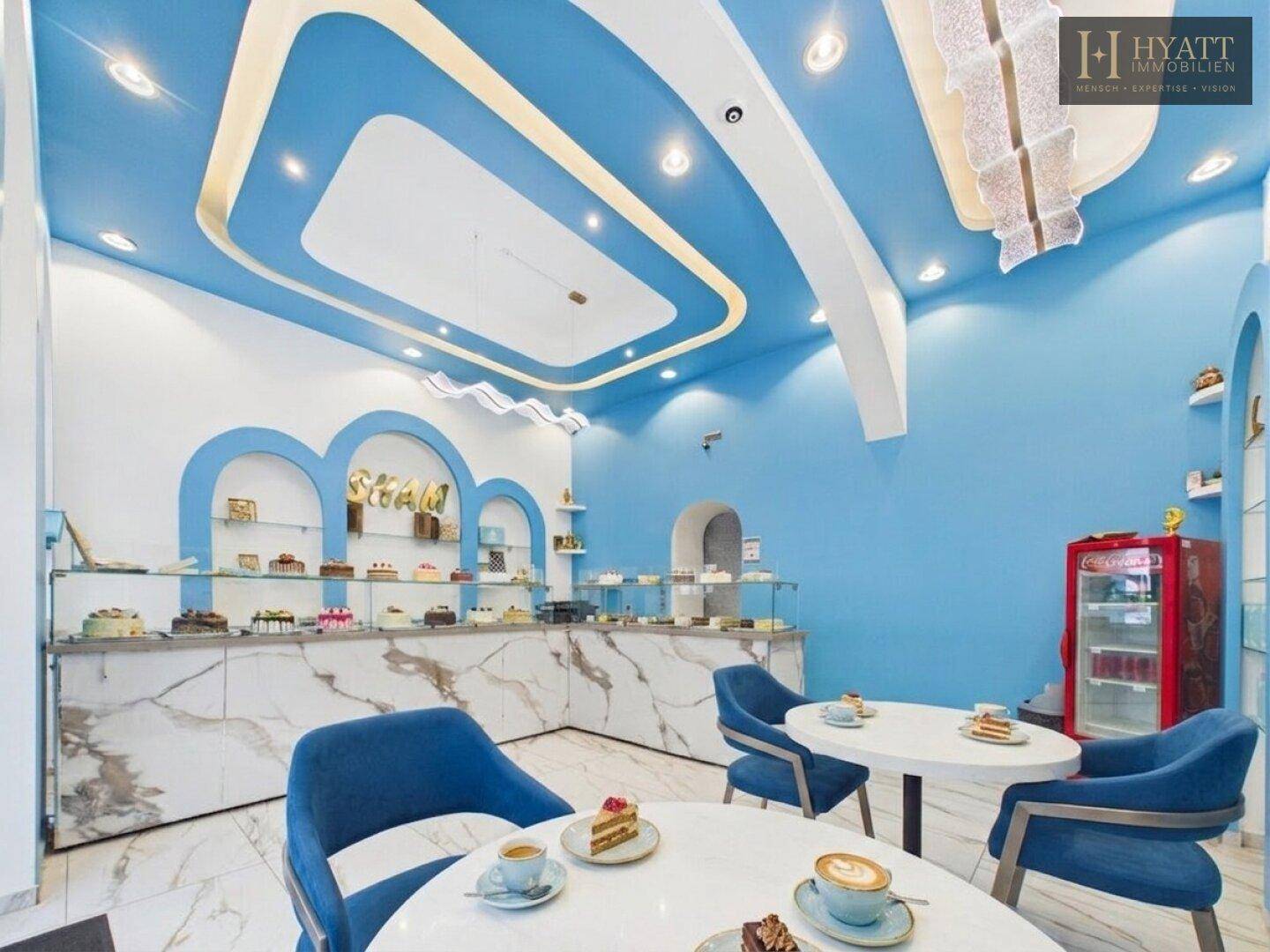 Gemütlicher Sitzbereich in einem Café mit blauen Stühlen, runden Tischen und einer Auswahl an Kuchen.