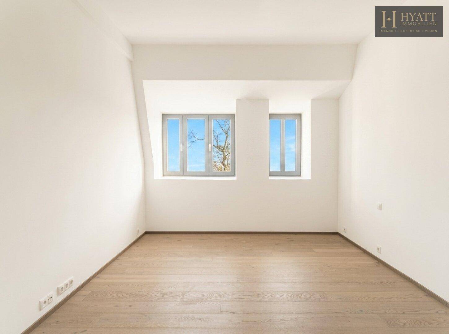 Helles Zimmer mit Holzboden, weißen Wänden und zwei Fenstern mit Blick ins Grüne.