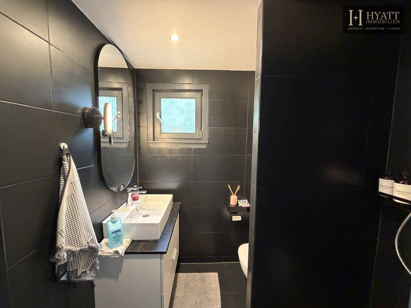 Modernes Badezimmer mit dunklen Fliesen, einem Waschbecken und einem ovalen Spiegel.