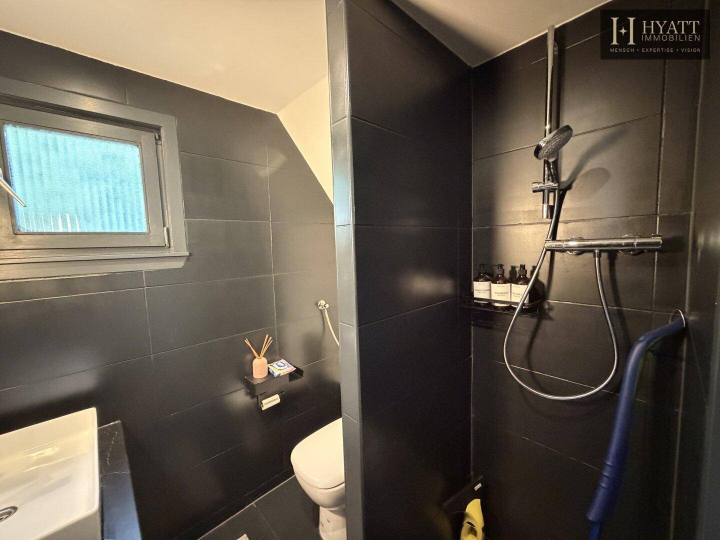 Dunkel gefliestes Badezimmer mit Dusche, Toilette und einem Fenster für natürliche Belüftung.