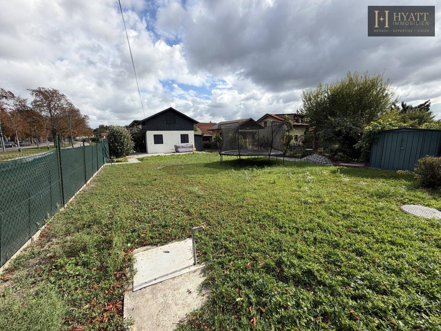 Gartenbereich mit Rasenfläche, Trampolin und einem Gartenschuppen unter bewölktem Himmel.