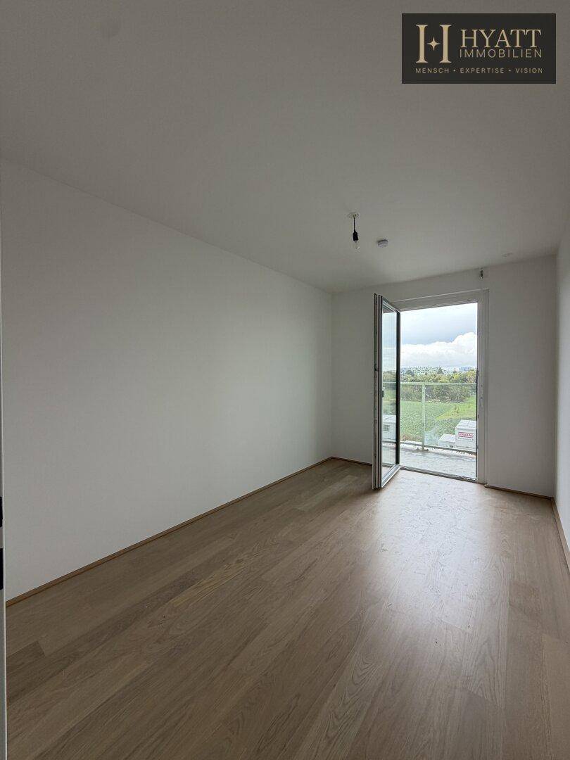 Leeres, helles Zimmer mit Holzboden und Zugang zu einem Balkon oder einer Terrasse.