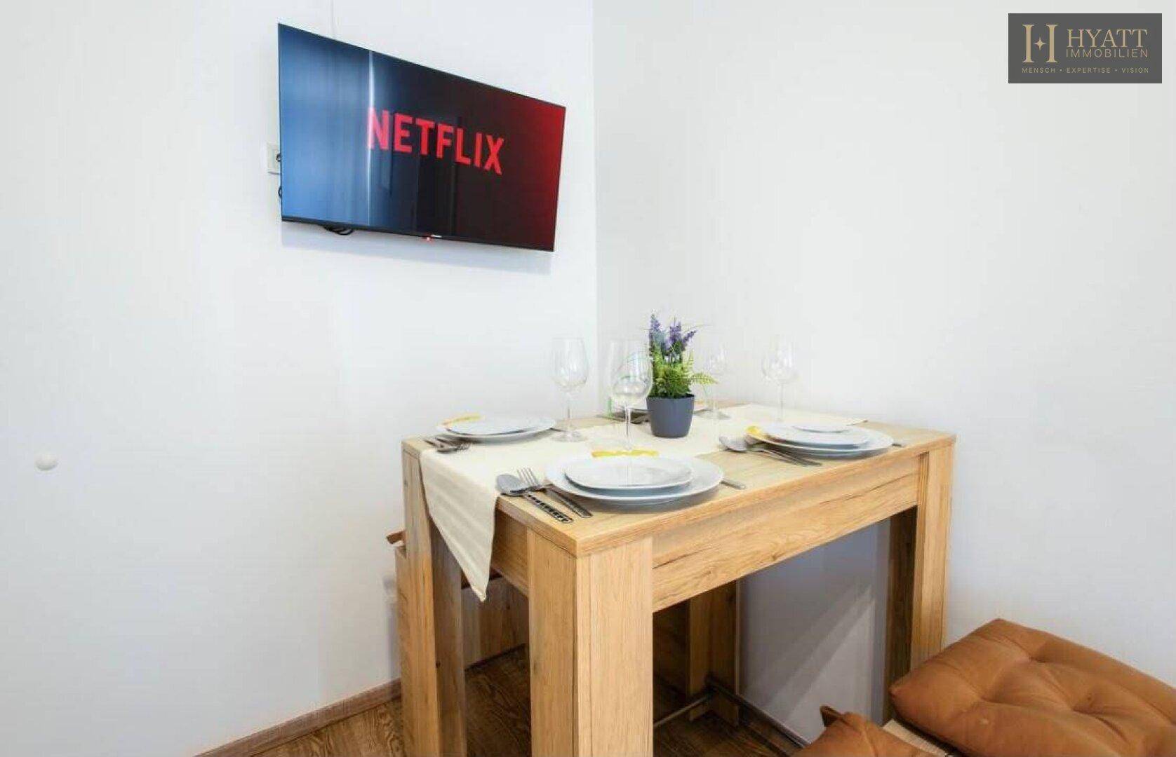 Kleiner Esstisch aus Holz, gedeckt für zwei Personen, mit Blick auf einen Wandfernseher.