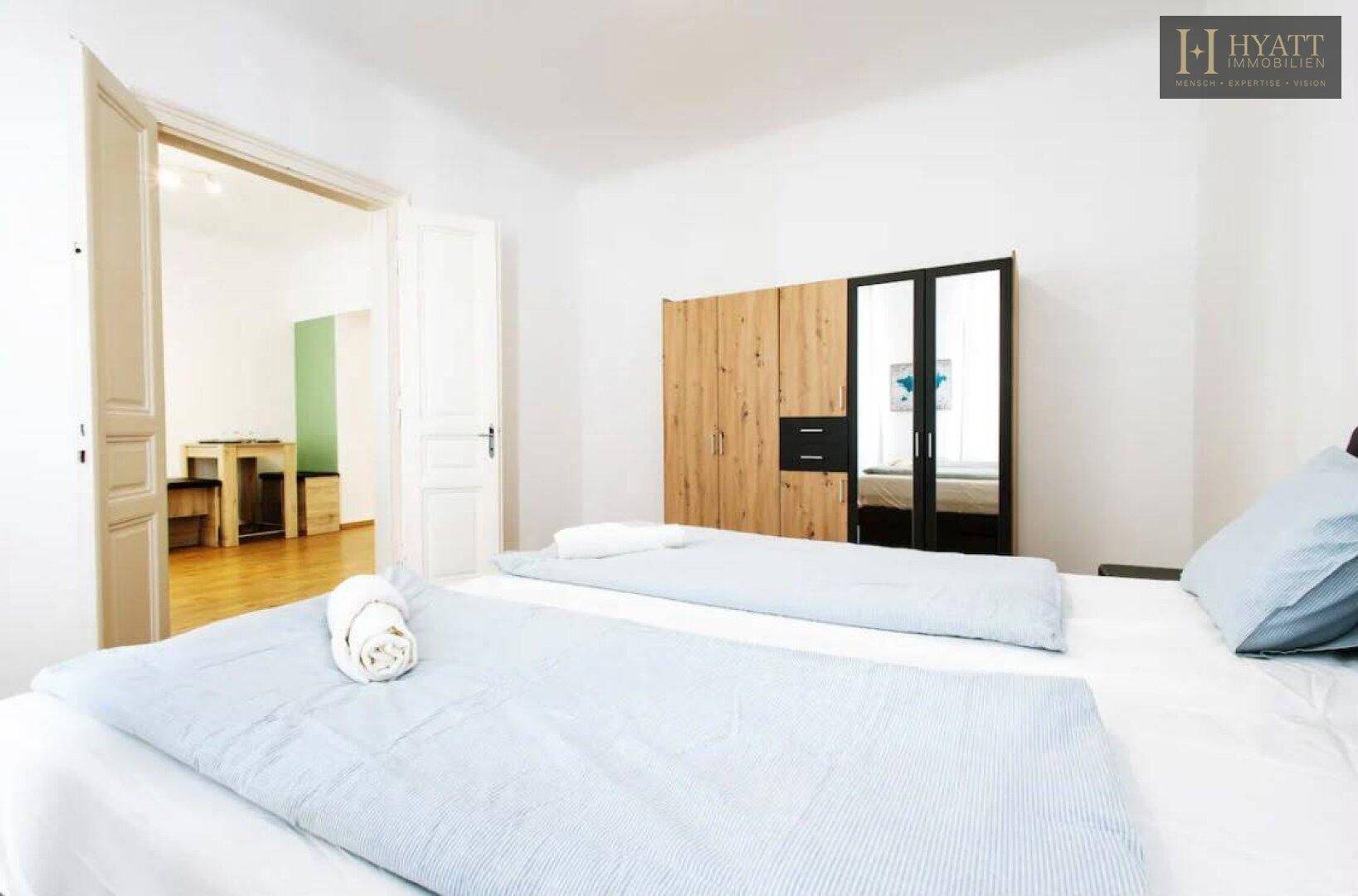 Schlafzimmer mit Doppelbett und Holzkleiderschrank, offen zum Wohnbereich hin.