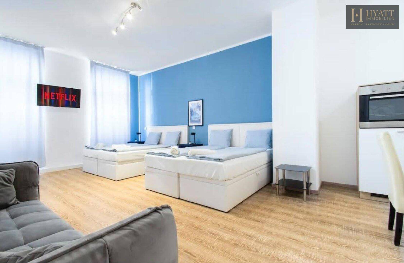 Helles Studio-Apartment mit zwei Betten, Sofa und blauer Akzentwand, inklusive kleiner Küchenzeile und Fernseher.