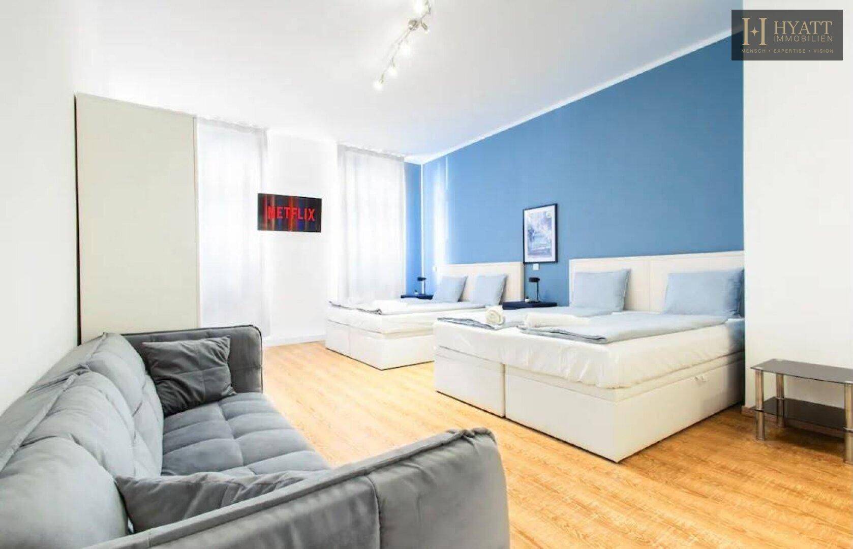 Geräumiges Studio-Apartment mit zwei Einzelbetten, bequemer Couch und einem Fernseher an der blauen Wand.