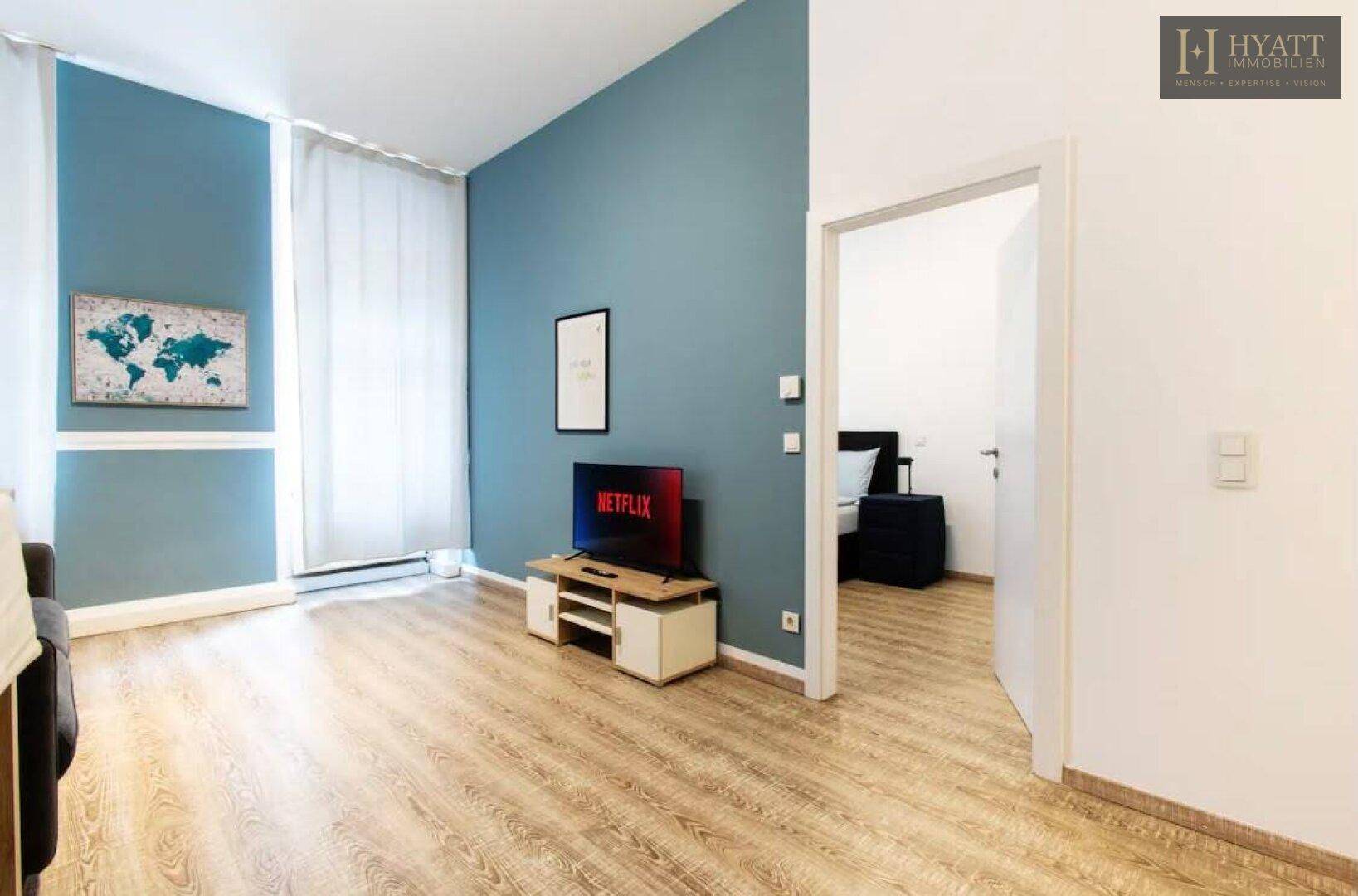 Heller Wohnbereich mit blauer Akzentwand, Sofa und Fernseher, angrenzend an ein weiteres Zimmer.
