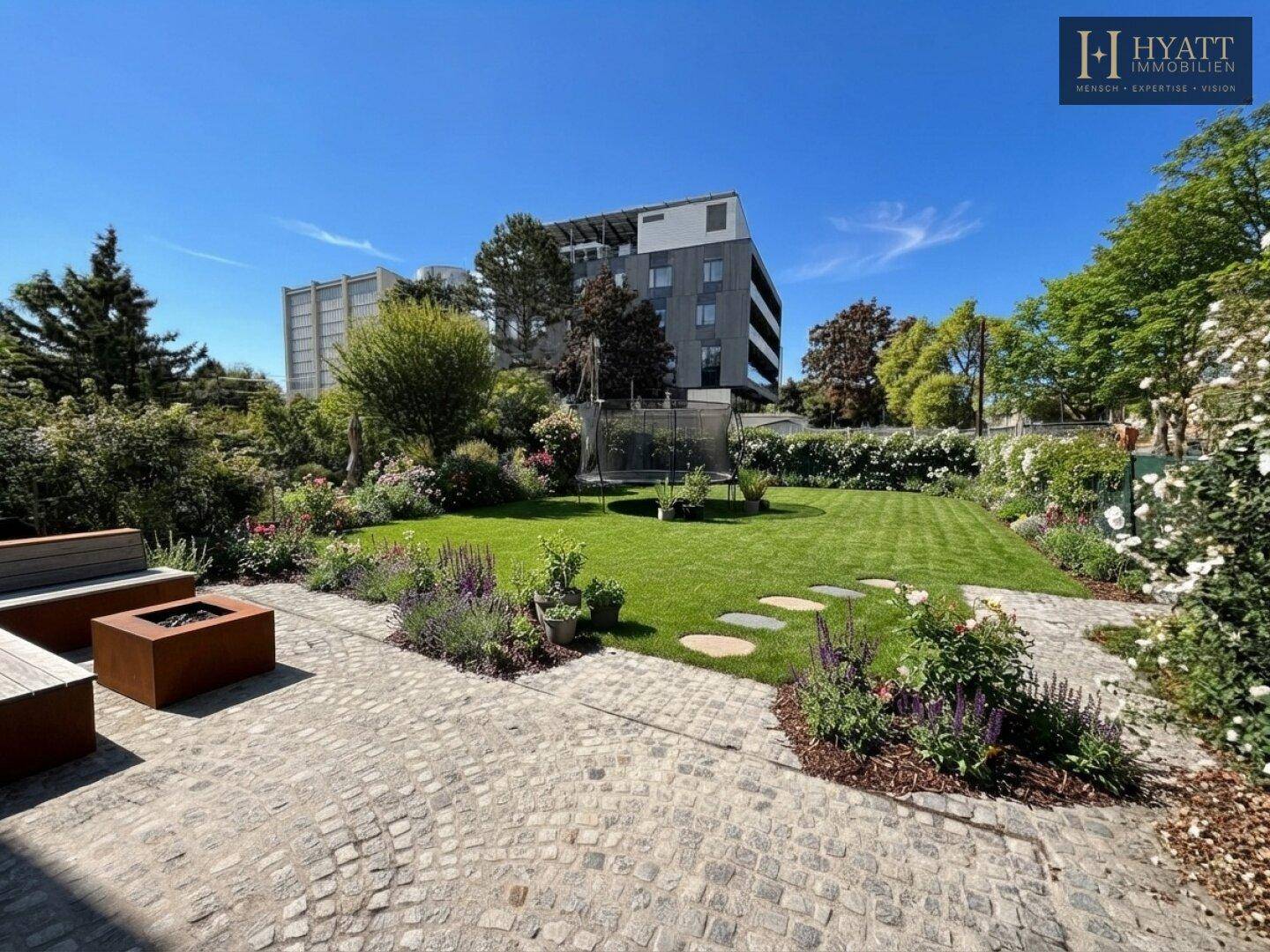 Weitläufiger Garten mit Rasenfläche, Trampolin und Blick auf ein modernes Gebäude im Hintergrund.
